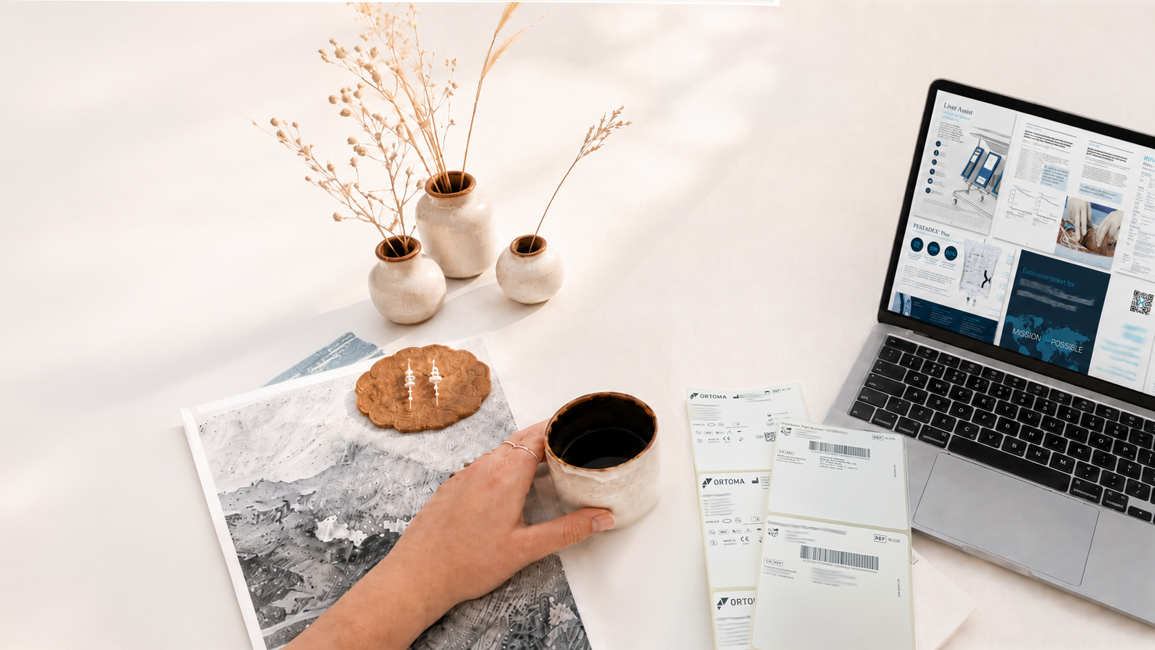 Studio still life with ceramics, printed labels and a laptop showing MedTech layouts.